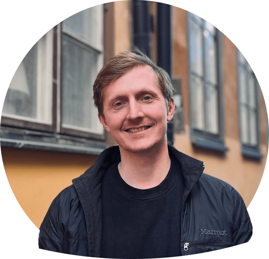 Santino ler mod kameraet foran en gul væg i Gamla stan, iklædd sort jacka og t-shirt.